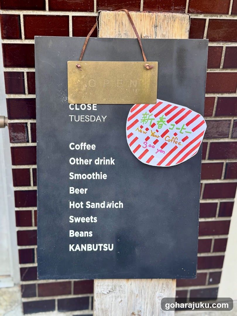 A black menu board hangs on a brick wall, displaying the shop's offerings and hours. The sign lists items like coffee, smoothies, beer, hot sandwiches, sweets, beans, and KANBUTSU. A 'CLOSE TUESDAY' notice is visible above the list. A red and white striped sticker with Japanese text and '300 yen' price tag is attached to the right side of the board.