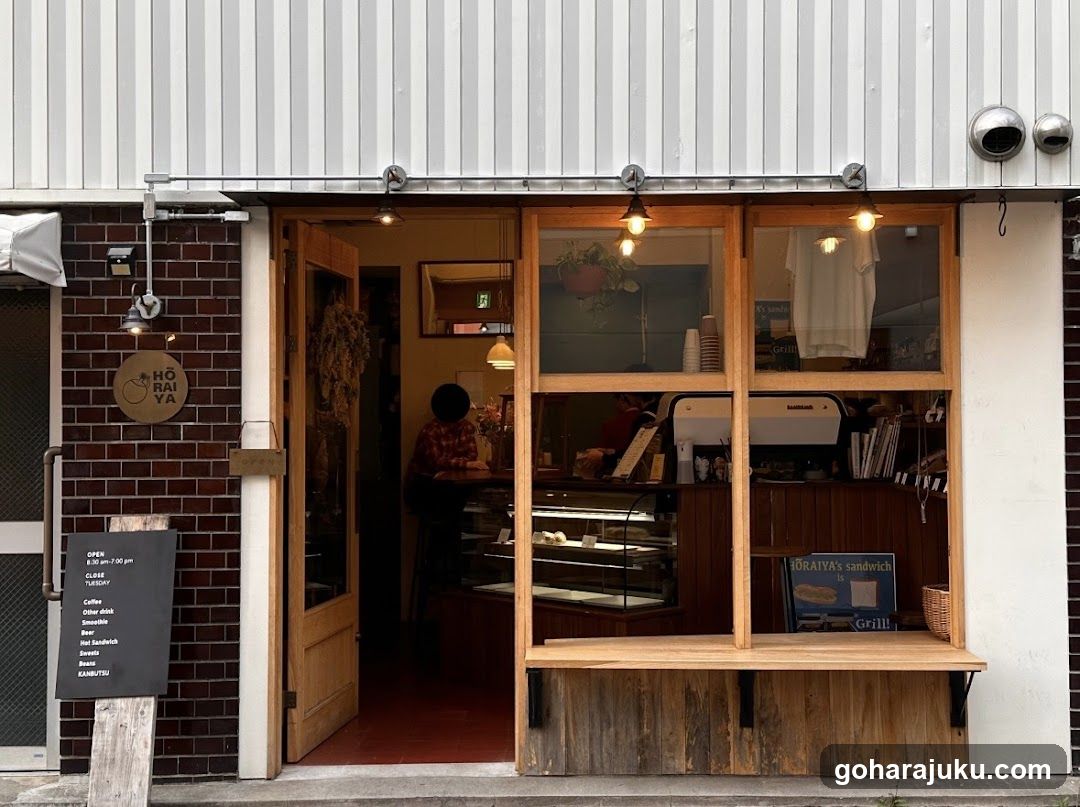 A small café or bakery with a wooden entrance and large display windows showing interior equipment, pastries in glass cases, and staff working behind the counter. A chalkboard menu stands outside listing coffee drinks and hours.