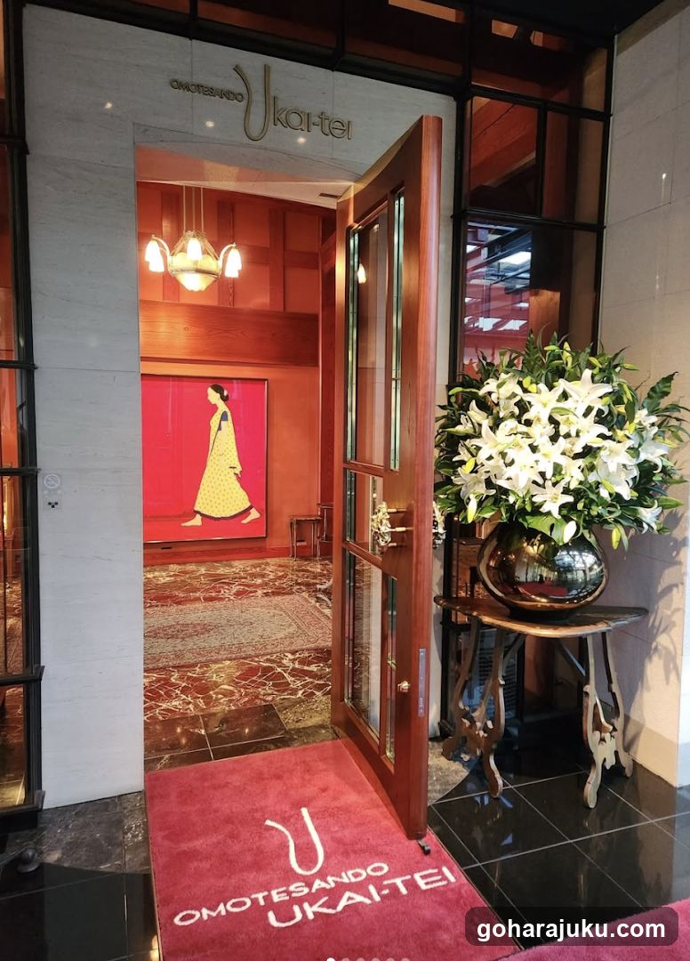The image shows the entrance of a building with 'OMOTESANDO UKAITEI' signage above the door. A red welcome mat with the same branding is placed at the doorway. To the right, there is a large floral arrangement in a metallic vase on an ornate stand. Inside, warm lighting illuminates a hallway with wooden paneling and a framed artwork depicting a figure in traditional attire.