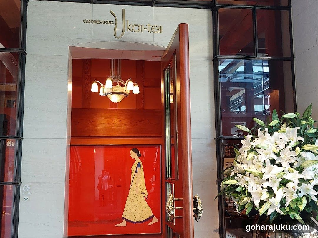 The image shows the entrance to a shop, likely a restaurant or cafe, featuring a sign that reads 'OMOTESANDO Ukai-tei'. The entrance is framed by large glass windows and doors, with a chandelier hanging inside. A vibrant red panel on the door displays an illustration of a woman in traditional attire. To the right, there is a lush arrangement of white lilies in a vase.