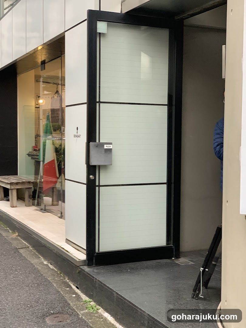 The image shows a modern storefront entrance featuring a glass door with frosted panels and black framing. A small sign is visible on the door, though its text is not legible. To the left, there is a window display showcasing various items, including what appears to be clothing or accessories. The building has a clean, contemporary design with light-colored walls and dark accents.