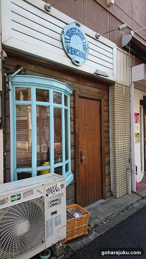 A quaint shop with a wooden facade, featuring a light blue bay window on the left and a brown door in the center. Above the entrance is a circular sign with text that reads 'BLUE CREAM KITCHEN'. An air conditioning unit sits outside near the ground, next to an orange crate.