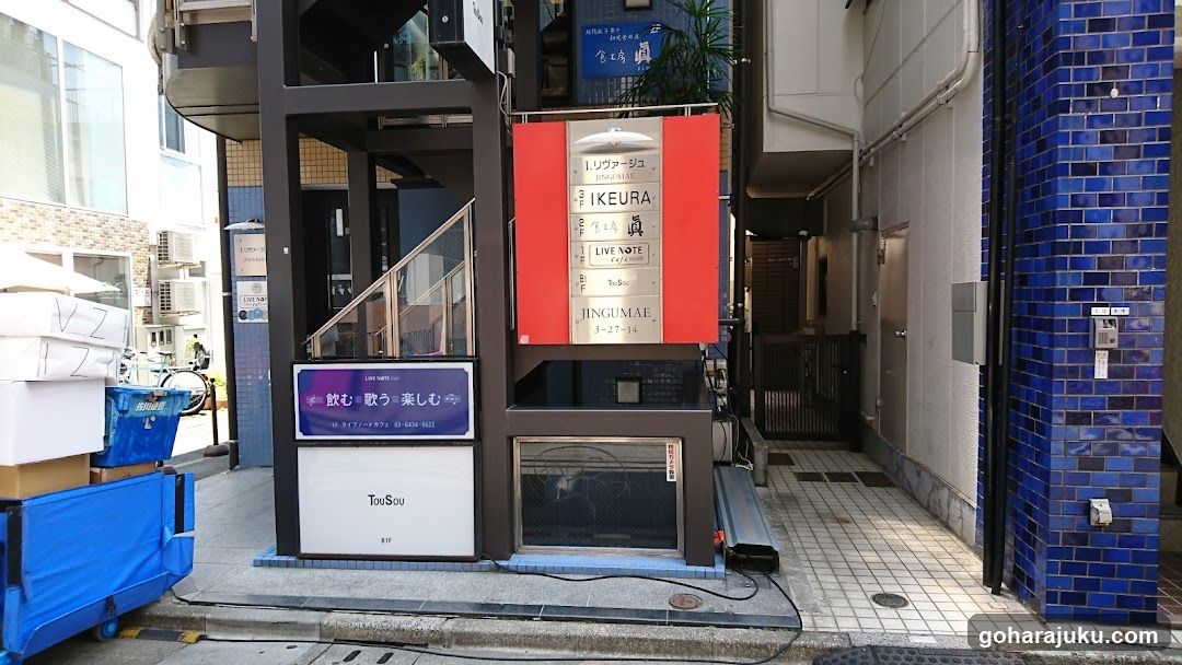 A modern building entrance with a glass elevator and signage reading 'IKEURA LIVE NOTE' in Japanese and English. A blue signboard displays promotional text, while stacked boxes and bins are visible nearby.