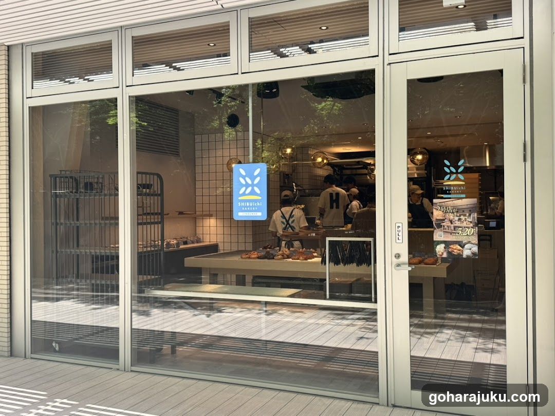 A modern bakery storefront with large glass windows and doors, displaying staff inside preparing food. A blue sign with a logo is visible on the window.