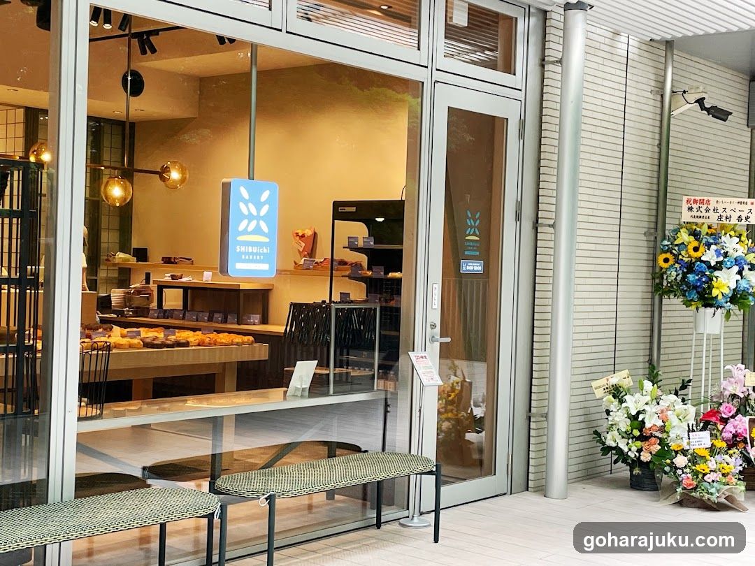 A modern bakery storefront featuring large glass windows and doors, displaying an array of baked goods inside. A blue illuminated sign with a leaf logo is mounted on the window. Outside, there are several floral arrangements placed near the entrance, including sunflowers and mixed flowers. The interior shows shelves stocked with pastries and breads, along with hanging light fixtures.