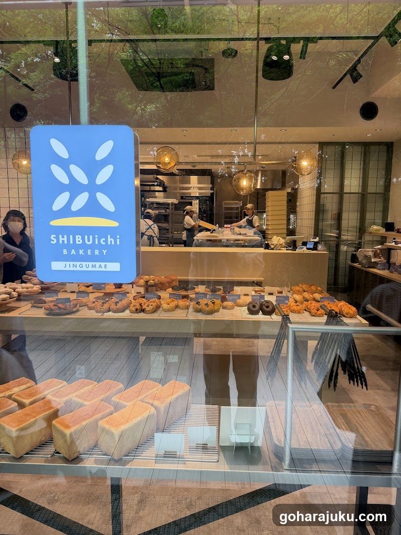 A photo of the physical exterior entrance/storefront facade of Shibuiichi Bakery Jingumae, with visible signage on the building. The display case showcases an array of baked goods including bread loaves and pastries.