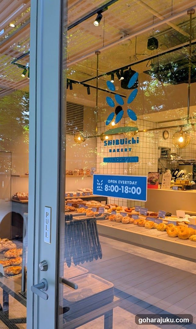 View through glass door showing interior bakery display with donuts and pastries on shelves, blue signage reading 'SHIBUICHI BAKERY' and 'OPEN EVERYDAY 8:00-18:00', pull handle visible on door frame.